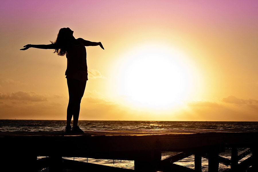 Trauma recovery shown by a woman with arms outstretched on dock at sunrise or sunset.