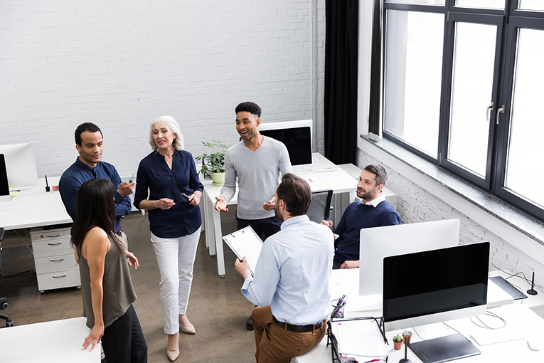 four men and two women meeting and talking together in a workplace.